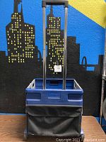 Front view of blue Staples foldable cart with black pouch and visible bottom crack.
