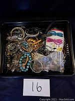 Top view of black plastic basket filled with various vintage costume jewellery pieces including beaded bracelets, metal chains, rings, earrings, and dragonfly hair clips on a card, presented with lot label '16'.