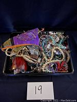 Photo showing a black tray filled with a mix of vintage costume jewelry pieces including necklaces, bracelets, brooches, and a purple pouch with embroidered detail.