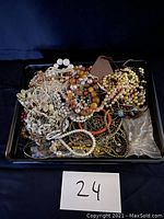 Overhead photo showing a black tray filled with tangled vintage costume jewelry consisting mostly of necklaces made from various beads and faux pearls, plus a pair of earrings and a small pouch. The photo includes a white card with the number 24.
