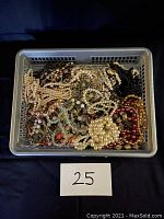 Photo showing a plastic basket filled with assorted vintage costume jewellery including many pearl-style necklaces, black and reddish beaded necklaces, clip-on earrings, and bracelets. A label with number '25' is visible.