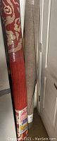 Photo showing two rolled rugs standing upright. One is red with beige ornamental patterns and the other is solid beige.