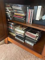 Photo showing stacks of CDs, DVDs, and VHS tapes placed in wooden cabinet, some plastic cases and boxes visible.
