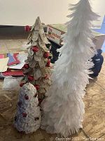 Three decorative Christmas trees pictured on floor; one is white and made of feathers, one is textured tan or beige with red ornaments, another is small and white with red accents.