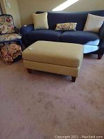 View of the ottoman in a living room setting near a blue sofa and patterned chair, showing fabric texture and overall shape.