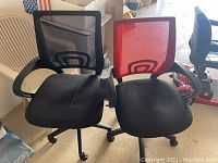 Two office desk chairs side by side; one black mesh back and one red mesh back, both with black seats and caster wheels. White plastic patio chair visible behind.
