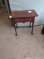 Photo showing one mahogany bedside table with a drawer and brass-tone handle. The table is on carpeted floor with a neutral background.