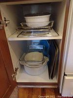 White CorningWare casserole dishes stacked inside a kitchen cabinet with clear glass lids on top, with glass Pyrex dishes and some metal sheet pans behind them.