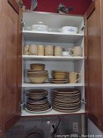 Cupboard with shelves holding multiple Thompson pottery dishes including plates, bowls, mugs, and a white Corning Ware baking dish with blue floral pattern on top shelf.