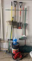Photo showing various yard tools including brooms, shovels, snow shovel, ice melt container, and a blue jug.