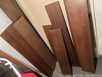 Seven mahogany shelving boards leaning against a wall in a storage area, showing wood grain and brown finish