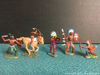 Five Britains American Wild West Native American toy soldier figurines posed in line on a green-blue carpet against a black backdrop. Includes standing warriors and one mounted on horse. Full set shown for condition and detail.