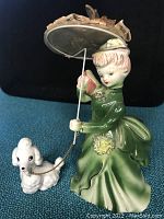 Side view showing woman in green dress holding parasol, porcelain dog on chain leash in front