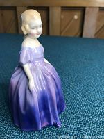 Porcelain figurine of a young woman in an ombre purple dress, showing side and front angles on blue textured surface.