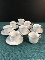 Photo of eight white porcelain cups each with gold rim trim placed on matching saucers against dark background