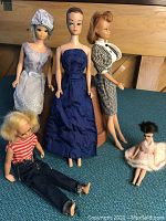 Five dolls arranged on green cloth: three full-size Barbie dolls standing behind two smaller companion dolls seated in front