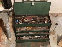 Open green metal toolbox with four drawers and top compartment showing assorted vintage hand tools including pliers, screwdrivers, wrenches, and files.