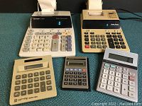 Five vintage desk top calculators on a blue surface, showing a variety of sizes and styles including paper roll models and handheld styles.