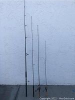 Four fishing rods standing against an exterior white wall, visible wear on handles and rods.