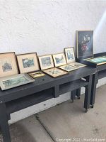 Nine framed artworks displayed on two tables under outdoor light