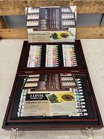 Closed and open views of 168 piece acrylic painting set with tubes of acrylic paint, brushes, pastels, and pencils in wooden case.