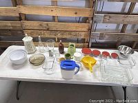 Wide shot of all kitchen items on a white table including bakeware, ceramic jar, mugs, mixing bowls, containers, and kitchen tools.