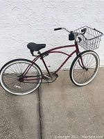 Full side view of maroon beach cruiser bicycle with black cushioned seat, front basket, and whitewall tires parked against a white stucco wall.