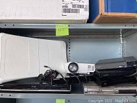 Shelf showing various electronic devices including DVD players, Blu-ray players, white projector, Wii Fit balance board, and FM transmitter.