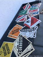 Overview photo showing a variety of hazard signs arranged on a table, including red, orange, black, white, and yellow signs with various hazard warnings.