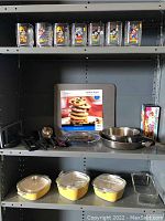 Shelf containing 13 Mickey Mouse themed glasses in varied designs on top shelf; middle shelf holds metal pans, utensils, a cookie sheet, and loaf pan in box; bottom shelf displays three yellow covered casserole dishes and a glass baking dish.