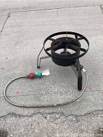 Overall view of large propane burner stand on concrete, showing metal circular burner with three legs and attached hose with regulator