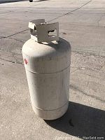 White metal propane tank standing on concrete with valve collar on top; shows surface wear and rust spots.