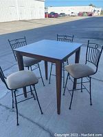 Photo showing the square wooden high top pub table and four wrought iron swivel chairs with upholstered seats outdoors.