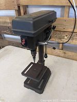Full view of small drill press showing metal construction, adjustable work table with slots, and step drill bit attached.