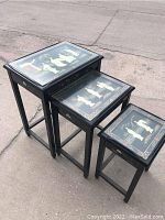 Three black lacquer nesting tables arranged from largest to smallest outdoors on pavement. Each table has a glass top with oriental mother of pearl inlay art visible beneath.