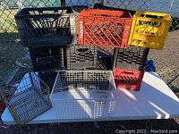 Assorted black, red, and yellow plastic milk crates stacked with two metal wire crates on a white table outside next to a chain link fence.