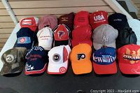Photo showing all 18 assorted baseball caps with visible logos including Budweiser, Atlanta Falcons, Honda Racing, Philadelphia Flyers, New York Rangers, New York Yankees, and an event commemorative cap.