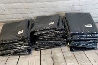 Stack of 29 black banquet chair covers, new and still sealed in packaging, resting on wooden floor against a white brick wall.