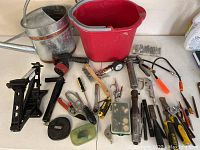 Mechanical car jack, watering can, red plastic bucket, pliers, caulking gun, tire gauge, screwdrivers, clamps and other assorted hand tools spread on a table