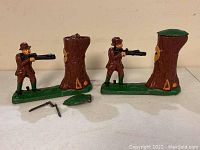 Two cast iron mechanical banks with hunter figures aiming rifles at tree stumps. One bank has loose pieces including a green top piece and a crank handle on the ground.