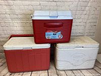 Photo showing all three coolers: one red Rubbermaid cooler on top of two white Coleman coolers stacked on a wooden floor with a white brick background.