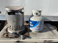 Photo showing a metal construction propane heater with ventilation slots and bent metal on a flat surface beside a Blue Rhino propane tank with valve.