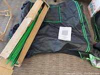 Grow tent black fabric with green trim, partially unrolled, green metal frame poles and connectors in cardboard box, manual on fabric.