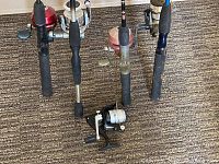 Four fishing rods standing vertically on a textured carpet, each fitted with a spinning reel attached, displaying foam handles and reel crank handles.