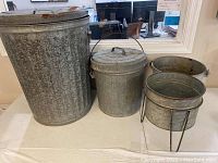 Set of four galvanized containers on table for scale: large trash can, smaller bucket with lid, bucket with handle, and planter with holder