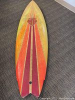 Full view of the Mark Richards surfboard showing the pointed nose, colorful stripe design, yellowed and worn surface, and fish tail shape with missing fins.