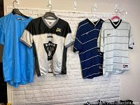 Four men's sports jerseys hanging on hangers; blue solid, black and silver Oakland Raiders NFL jersey, blue striped Nike jersey, and white striped Nike jersey
