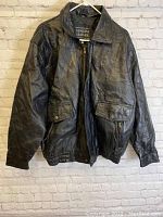 Front view of men’s black leather jacket hung on hanger against brick wall background showing collar, zipper closure, and flap pockets with snaps.