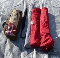 All items shown laid on outdoor tarp: Quest screen house bag, 2 red folding chairs in fabric bags, grey and black collapsible umbrellas.