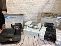 Three printers displayed side by side: HP OfficeJet 4652 in its box, white HP LaserJet multifunction printer, and Canon PIXMA MG6820 with packaging foam and box in background.